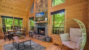L’intérieur chaleureux d’un chalet avec une cheminée en pierre, des murs en bois et de grandes fenêtres donnant sur la forêt.