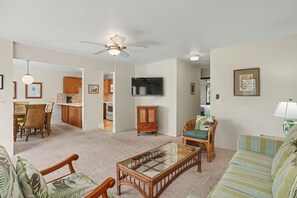 A spacious living room with a glass coffee table, a television, and a dining area with wooden cabinets and a tiled backsplash.