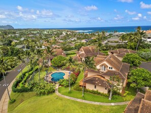 A residential area with a large swimming pool surrounded by lush greenery and palm trees. Houses with brown roofs and white walls are visible.