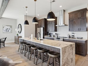 A modern kitchen with a large island, bar stools, dark wooden cabinets, and pendant lighting.