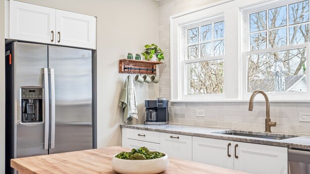 Una cocina moderna con armarios blancos, un refrigerador de acero inoxidable, una cafetera y una ventana con vista a árboles.