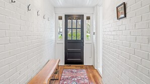 Un pasillo con paredes de ladrillo blanco, un banco de madera, una puerta negra con ventanas y un candelabro colgante.