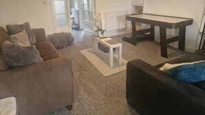 Ein Wohnzimmer mit einer braunen Couch, einem schwarzen Hocker, einem weißen Couchtisch und einem schwarzen Tisch. Eine Tür führt in einen anderen Raum, und an der Wand ist eine Heizung angebracht.