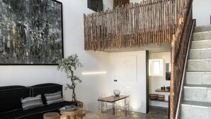 Uma sala de estar moderna com um sofá preto, cadeiras transparentes, uma pequena mesa de madeira, um grande quadro abstrato e uma escada com corrimão de madeira.