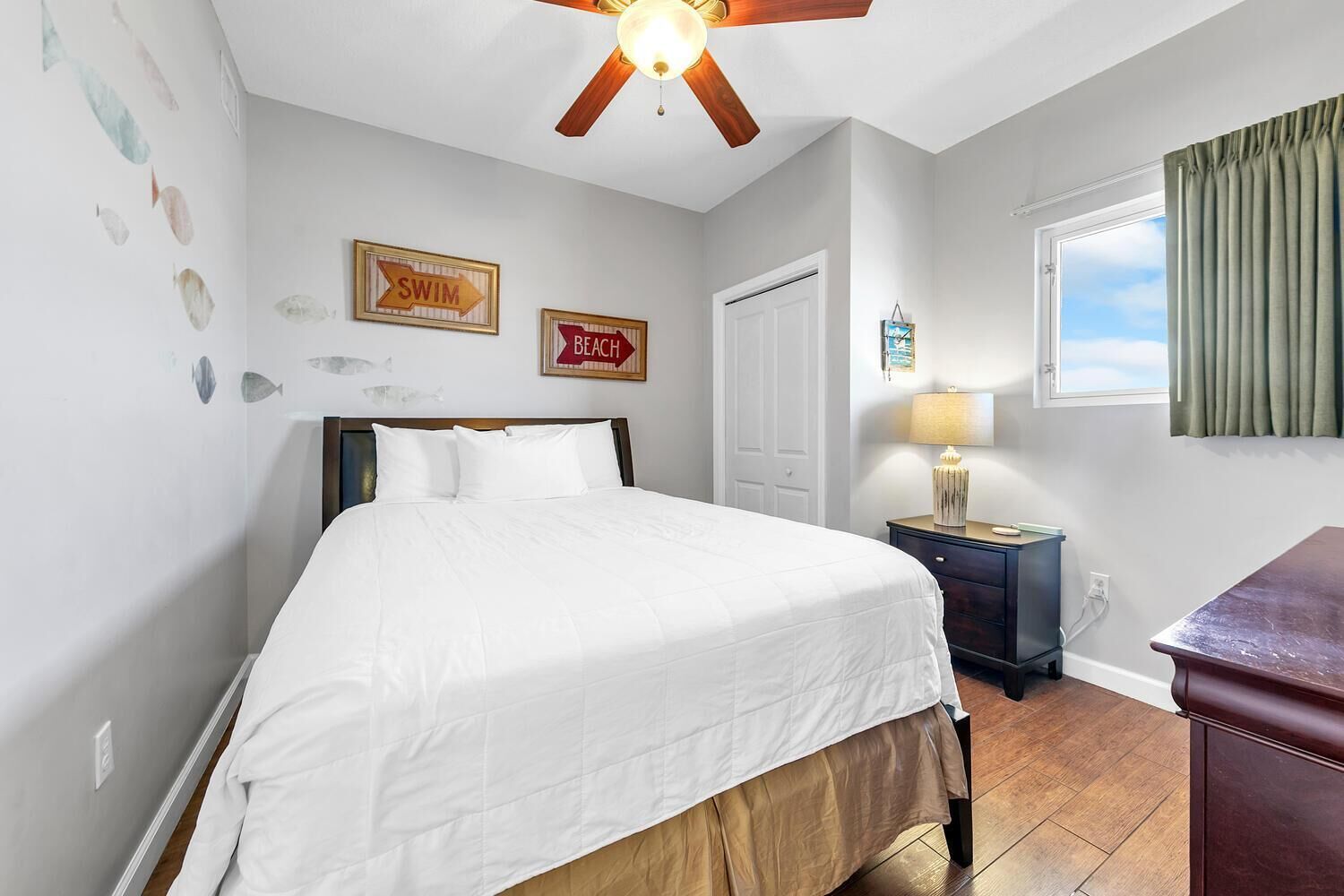 A bedroom with a bed, a nightstand, a ceiling fan, a window with curtains, and wall decorations including fish and directional signs.