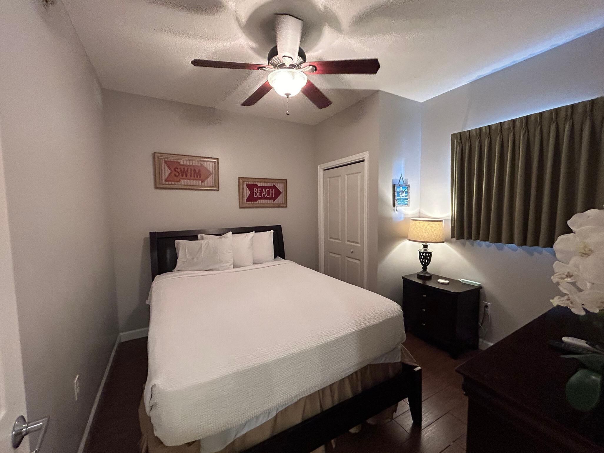 Une chambre d’hôtel avec un lit, une table de chevet, une lampe, un ventilateur de plafond et deux panneaux accrochés au mur indiquant les mots « PISCINE » et « PLAGE ».