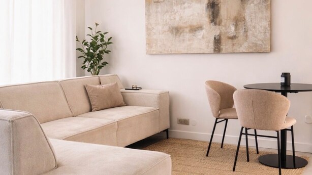 A modern living room with a beige sectional sofa, a small round table, two chairs, a large abstract painting, and a potted plant. - Maison Calme en Bordure de Forêt (Le Perray-en-Yvelines)