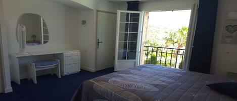 Een slaapkamer met een bed, bureau, stoel, kledingkast en een balkon met uitzicht op groen.