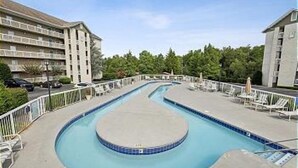 A swimming pool with a curved design, surrounded by lounge chairs and umbrellas, with buildings in the background. - Walk-in Level | 2 BR | Balcony | Sleeps 6 | Free Tickets (Pigeon Forge)