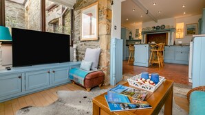 A cozy living room with a TV, seating, and magazines. A kitchen with blue cabinets and a dining area with stools is visible in the background. - 1 bedroom accommodation in Newquay (Newquay)