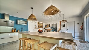 A modern kitchen with a large island, bar stools, hanging wicker chandeliers, and a blue accent wall. - Casa Marcel, 4 Chambres, 4 Sdb, Décoration Bohème, au Calme Dans la Pinède (La Tremblade)