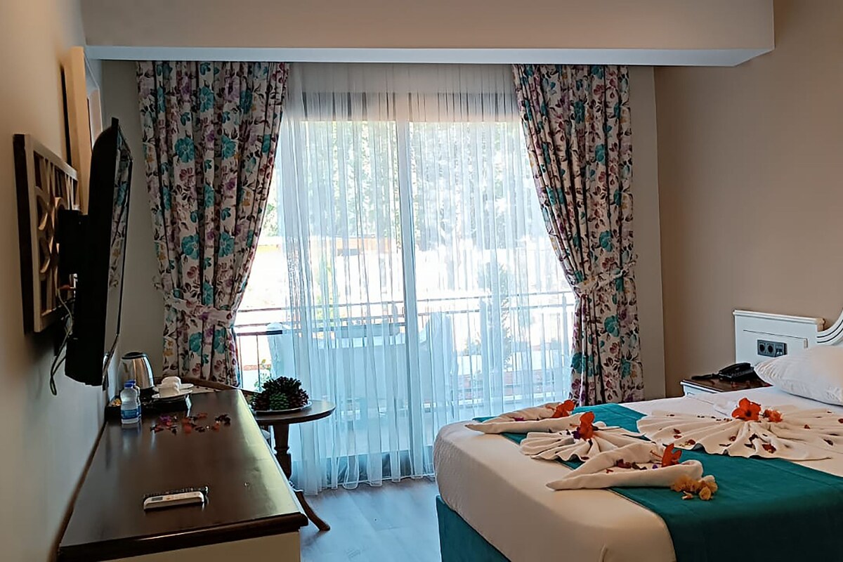 Una habitación de hotel con cama, escritorio, televisión y ventana con cortinas.
