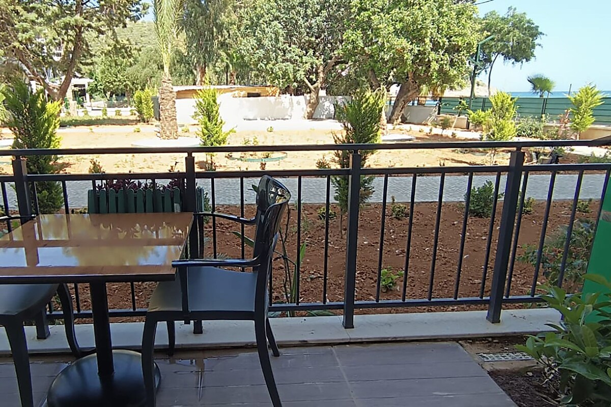 En balkon med et bord og stole, der giver udsigt over et sandet område med træer og hav.