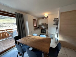 Eine kompakte Küche mit weißen Schränken, einem Holztisch und Stühlen. Vom Fenster aus ist ein Balkon mit Blick auf ein Feld zu sehen.