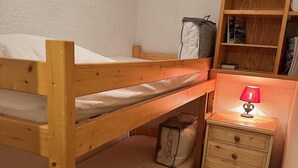 Kerrossänky, jossa on puinen pääty, yöpöytä lampun kanssa ja pieni kirjahylly.