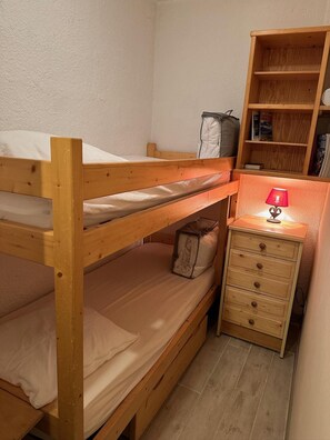 A bunk bed with a bedside table and a lamp, a small shelf with books, and a bag on the floor.