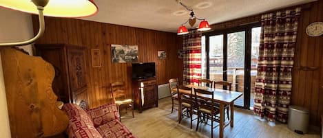 A cozy living room with a sofa, a television, a dining table with chairs, and a window with curtains.