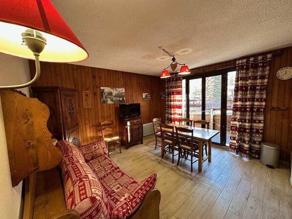 A cozy living room with a sofa, a television, a dining table with chairs, and a window with curtains.