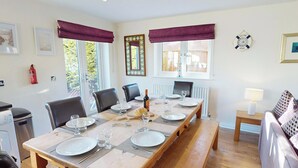 A dining area with a long wooden table set for four, surrounded by chairs, with a view of the outdoors through large windows. - Discover coastal bliss at The Dunes, your perfect family haven by Beadnell Bay. (Beadnell)