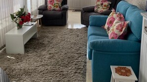 Una sala de estar con un sofá azul, dos sillones, una mesa de centro blanca y un taburete pequeño. Se aprecia un amplio ventanal con persianas, un ventilador de techo y un jarrón decorativo con flores sobre una mesita auxiliar.