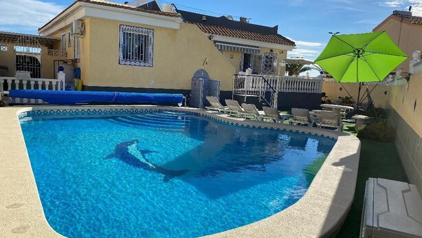 Una piscina de borde curvo, rodeada por una terraza color beige. Se observan sillas de descanso y un paraguas verde.