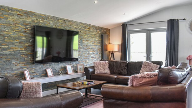 Un salon moderne avec des canapés en cuir, un téléviseur à écran plat fixé à un mur en briques, une table basse et une grande fenêtre avec des rideaux.