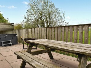 A wooden picnic table and benches on a patio with a wooden fence and a hot tub in the background.