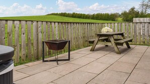 Une terrasse avec une table en bois et une brouette, entourée d’une clôture en bois et, au fond, une colline couverte d’herbe.