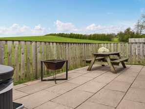 A patio with a wooden table and a wheelbarrow, surrounded by a wooden fence and a grassy hill in the background. - 4 bedroom accommodation in Kirkby Lonsdale (Kirkby Lonsdale)