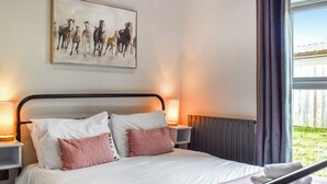 Un lit bien fait, avec des oreillers blancs et roses, une couverture blanche et une serviette enroulée posée sur une tête de lit en métal. On y trouve deux lampes de chevet, un tableau représentant des chevaux accroché au mur et une fenêtre avec des rideaux.