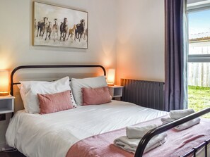 A neatly made bed with white and pink pillows, a white blanket, and a rolled towel on a metal headboard. There are two bedside lamps, a painting of horses on the wall, and a window with curtains. - 4 bedroom accommodation in Kirkby Lonsdale (Kirkby Lonsdale)