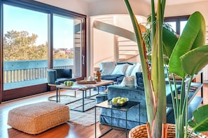 Uma sala de estar moderna com sofá, mesa de centro e um banquinho, uma escada e janelas amplas com vista para árvores.