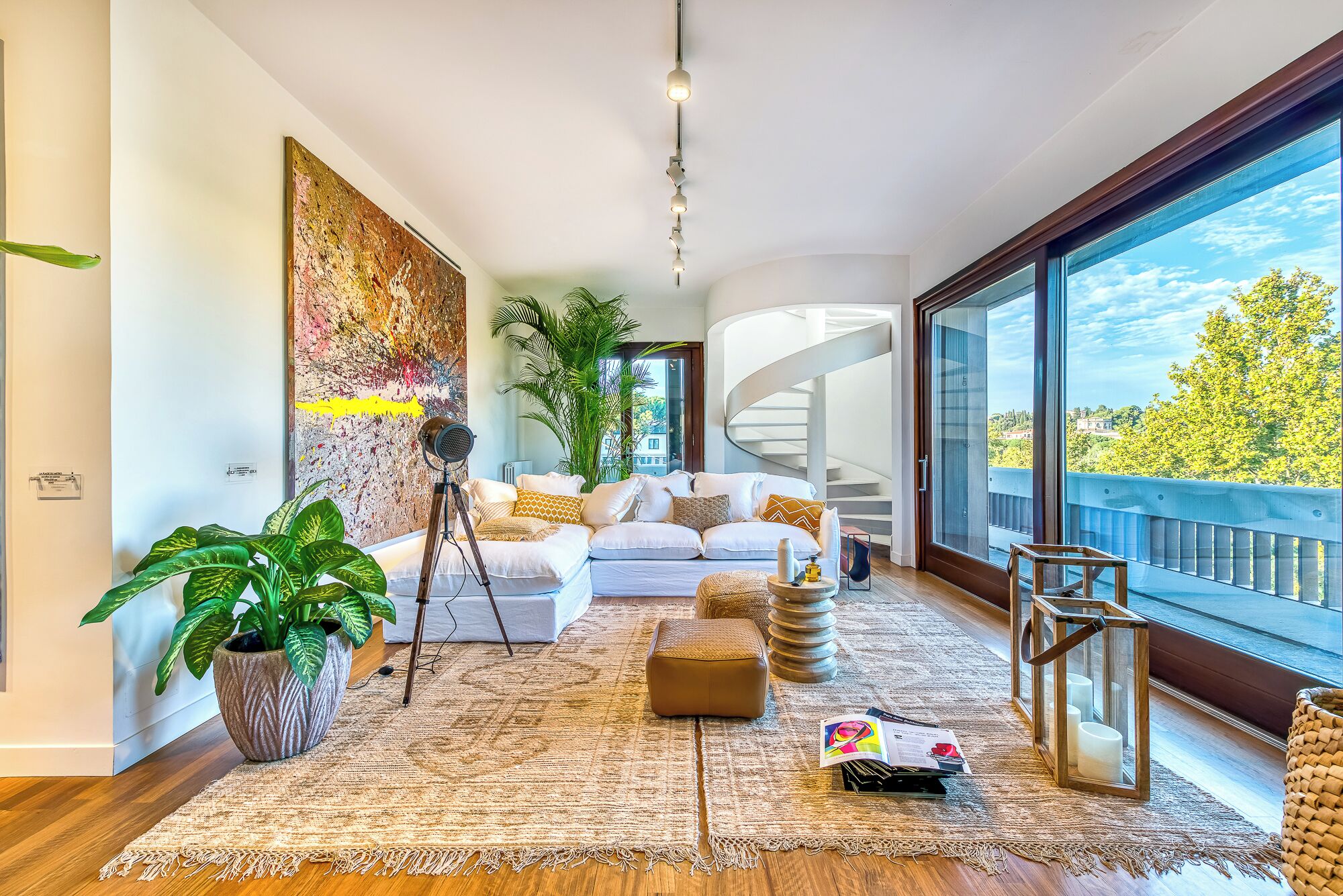 A modern living room with a white sofa, a large potted plant, a tripod, and a spiral staircase.