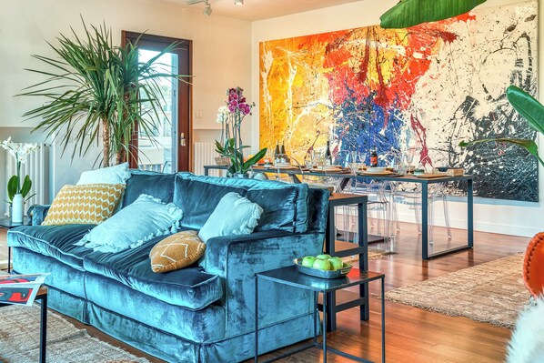 A modern living room with a blue sofa, a dining table, and a large abstract painting on the wall.