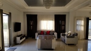A spacious living room with a large sofa, a smaller armchair, a flat-screen TV, and a chandelier.