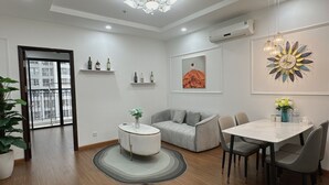 Una sala de estar moderna con un sofá gris, una mesa de centro redonda y un comedor con una mesa blanca y sillas grises. Hay un ventanal con vistas a edificios, y la habitación tiene una araña de techo y aire acondicionado instalado en la pared.