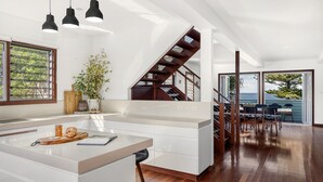 A modern kitchen with a central island, wooden staircase, and a dining area with a view of the ocean. - Allawah Cottage - Historic Beachside Retreat With Pippi Beach Views - Holiday Yamba (Yamba)