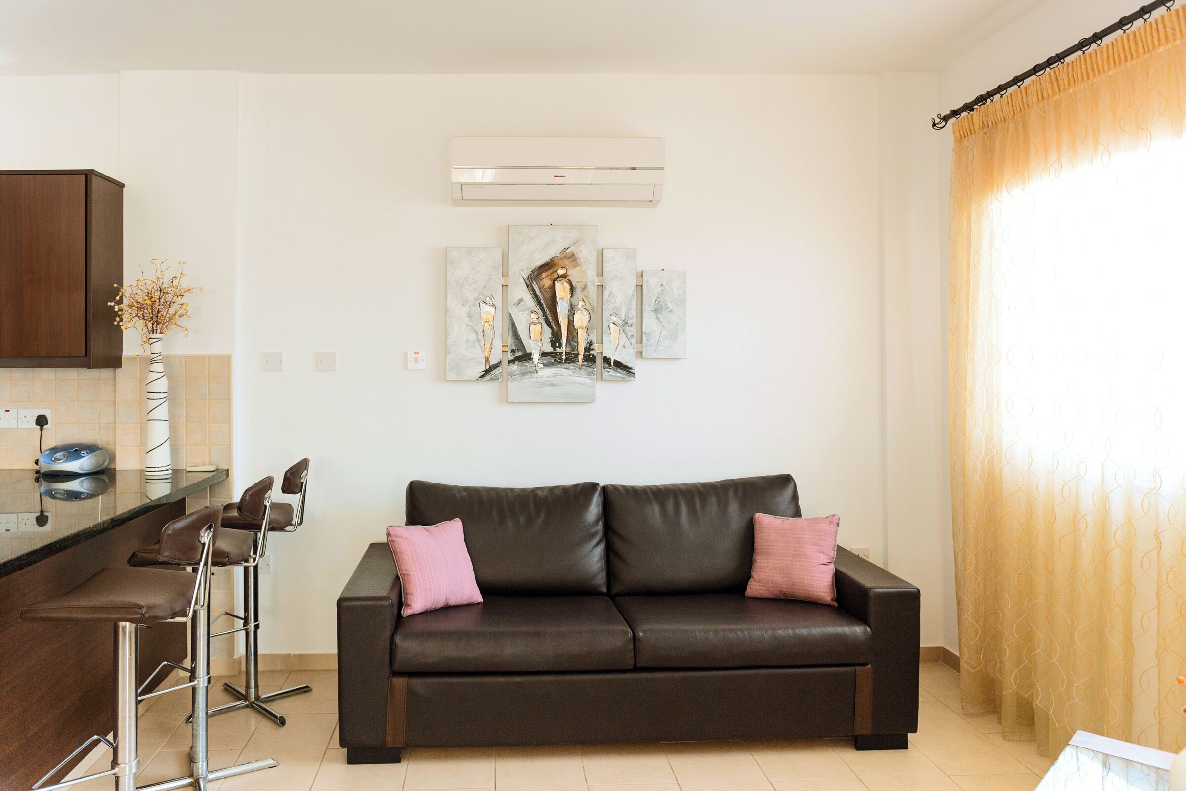 A modern living room with a dark brown leather sofa, two pink cushions, a coffee table, and a kitchen area with a bar stool and a vase of dried flowers.