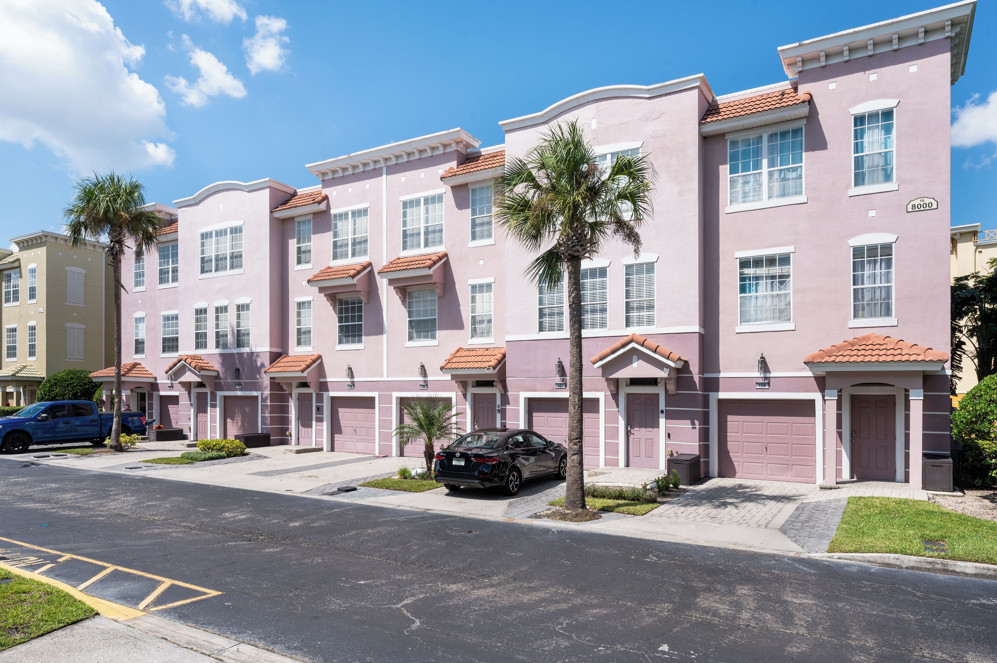 A row of pink townhouses with red-tiled roofs, a black car parked in front, and a blue truck on the left.