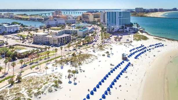 Eine Vogelperspektive auf eine Küstenhotelanlage mit mehreren Gebäuden, einem Strand mit Sonnenschirmen und einer Brücke im Hintergrund.