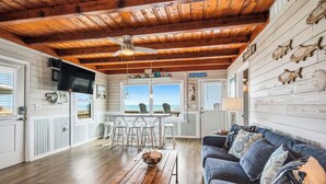A cabin with a wooden ceiling, a dining area with a view, a sofa, and a table with a bowl. - Dreamy Days: Coastal Cottage with Awe-inspiring Oceanfront Setting (Oak Island)