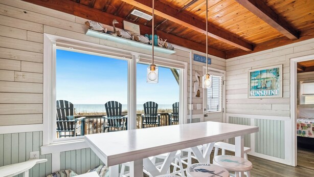 A beachside room with a white table, bar stools, and a view of the beach. - Dreamy Days: Coastal Cottage with Awe-inspiring Oceanfront Setting (Oak Island)