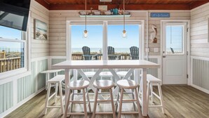 A room with a wooden ceiling, a white table with four stools, a door with a sign, and a view of the beach through large windows. - Dreamy Days: Coastal Cottage with Awe-inspiring Oceanfront Setting (Oak Island)