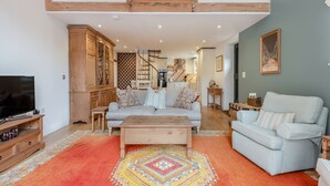 A spacious living room with a high ceiling, wooden beams, a staircase, a television, a sofa, a coffee table, and a patterned rug. - 1 bedroom accommodation in Broad Chalke (Broad Chalke)