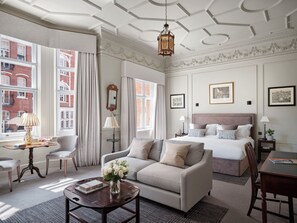 A hotel room with a large bed, a sofa, a coffee table, and a desk. There are two windows with curtains, a chandelier, and framed pictures on the walls. - The Chelsea Townhouse (London)
