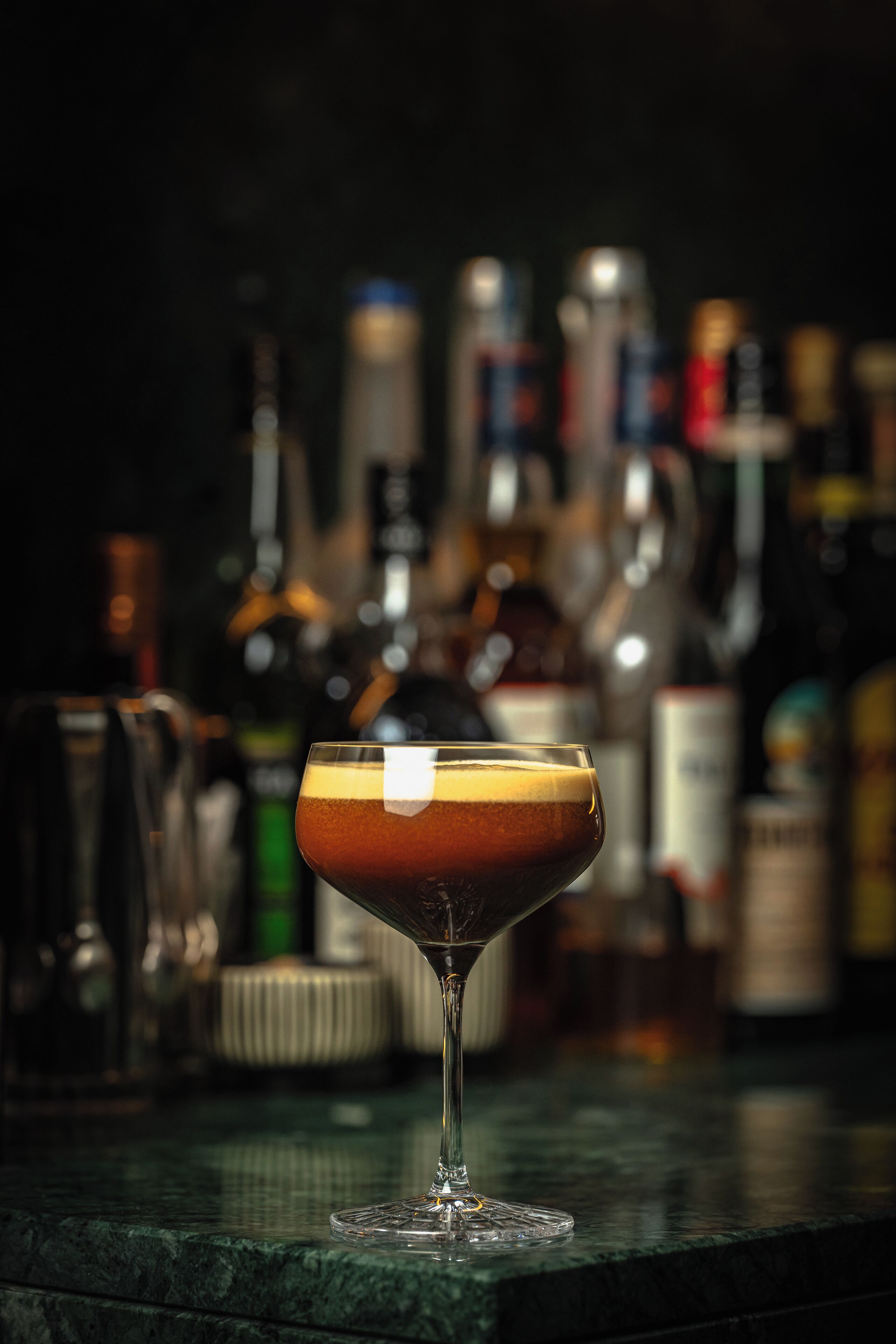 A cocktail in a martini glass on a bar counter with blurred bottles in the background.