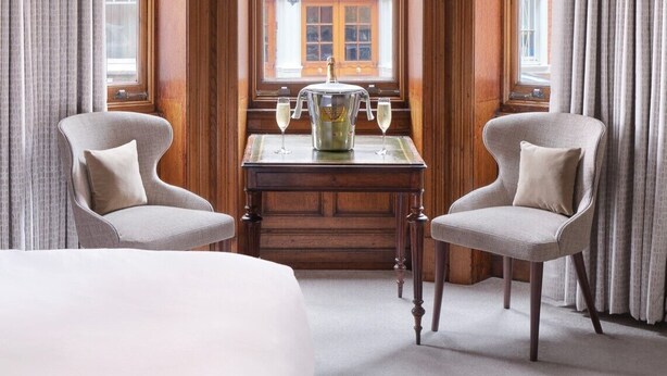 Habitación de hotel con un ventanal grande, una araña de cristal, dos sillones, una mesa con jarrón y vasos, y una cama.