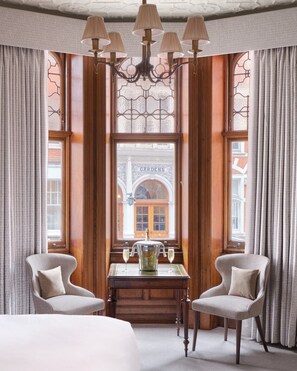 A hotel room with a large window, a chandelier, two armchairs, a table with a pitcher and glasses, and a bed. - The Chelsea Townhouse (London)