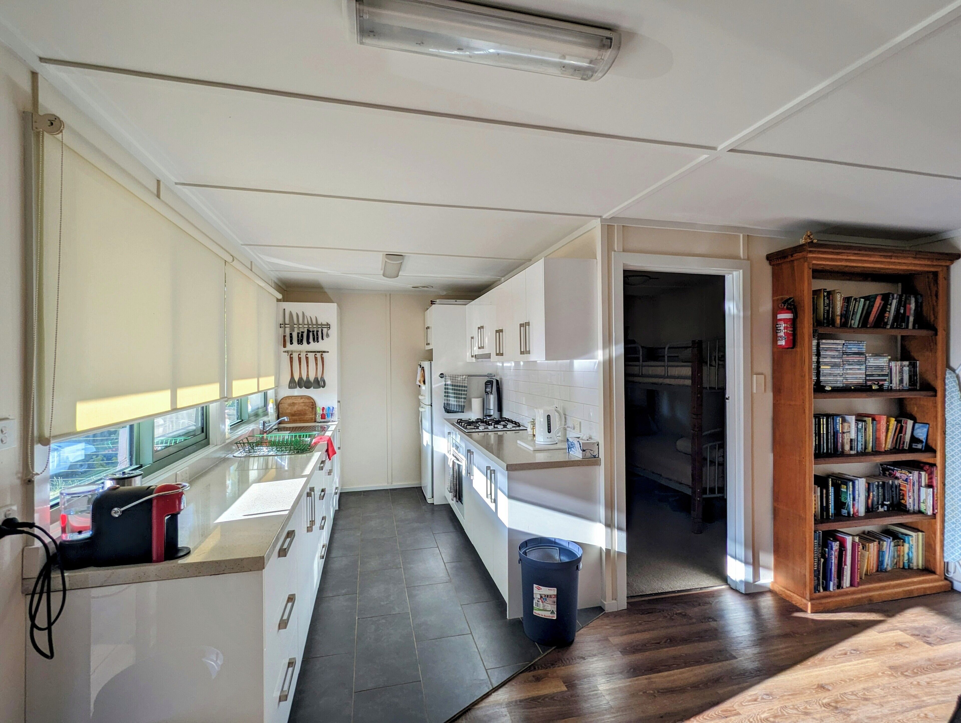 Une cuisine compacte avec des meubles blancs, une cuisinière encastrée et une machine à café. Une bibliothèque remplie de livres se trouve sur le côté droit.