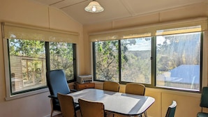A dining area with a table and chairs, large windows with views of trees and a building, and a ceiling light fixture. - Oak Cottage - Daylesford - 200m from everything (Daylesford)